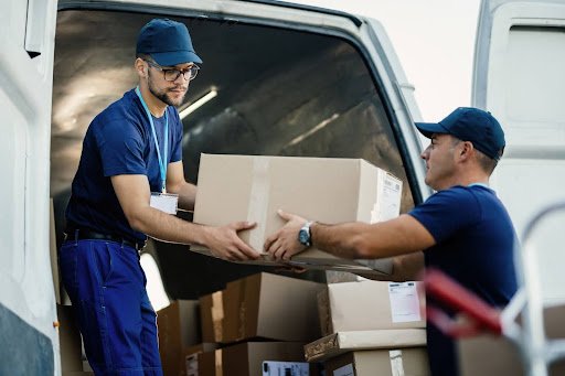 Logística reversa: Dois jovens trabalhadores de entrega em uniformes azuis descarregando caixas de uma van.