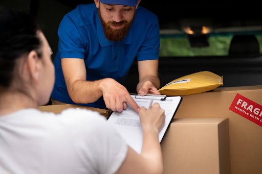 Diferencial da reversa: Um homem de meia-idade, com barba e cabelo castanho curto, vestindo uma camiseta azul, está na traseira de uma van repleta de várias caixas de papelão. Ele entrega uma prancheta para uma mulher de cabelo castanho, que está prestes a assinar um documento.