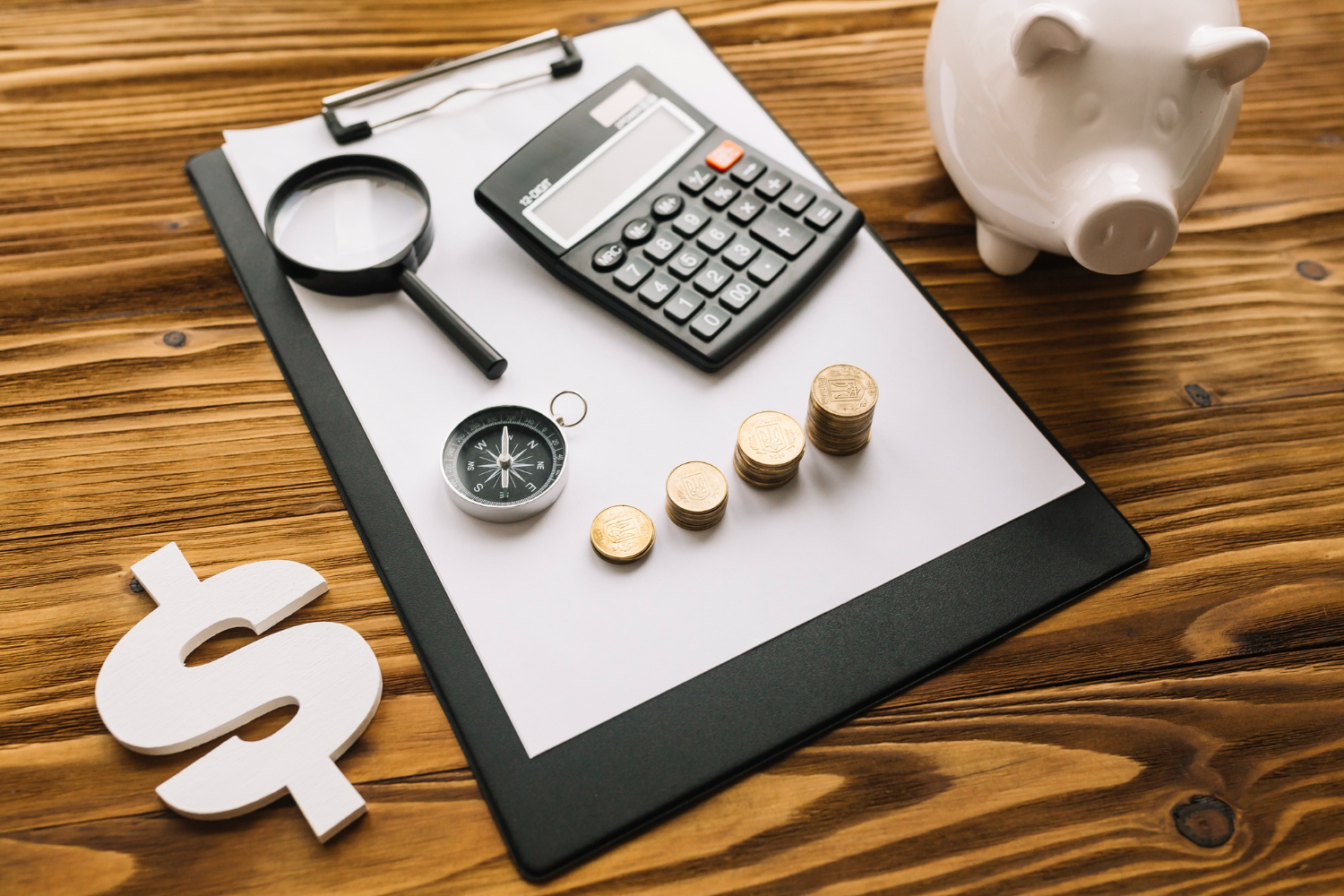 Eficiência operacional na Logística Reversa: Uma mesa de madeira com uma prancheta contendo moedas, uma lupa, uma calculadora e uma bússola. Na frente, há um sinal de cifrão. Ao fundo, um cofrinho de porquinho. A composição sugere planejamento financeiro.