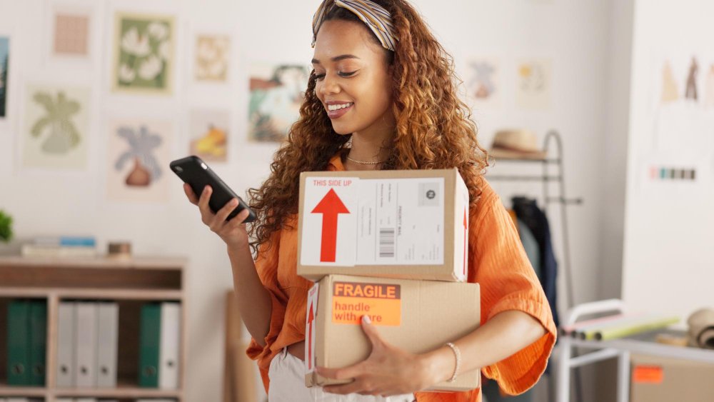 Recompra e fidelização: Uma jovem mulher com cabelo longo e cacheado castanho, vestindo uma camisa laranja, segurando uma caixa de papelão com um rótulo de 'Frágil', sorrindo e olhando para seu telefone.