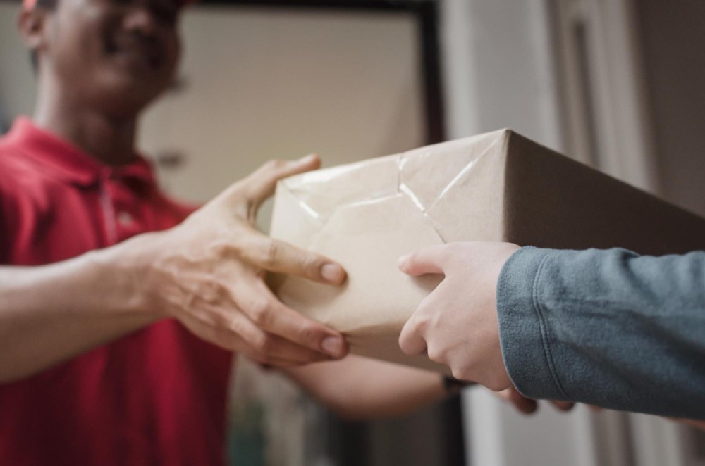 A Logística Reversa pode ajudar a sua empresa a crescer: Uma pessoa com uma camisa vermelha entregando uma caixa de papelão para outra pessoa com uma camisa azul.